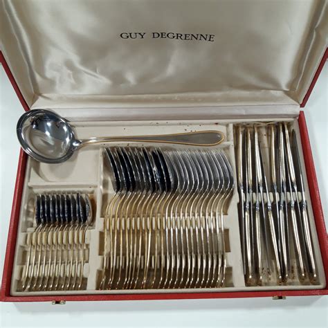 Photographie d'une ménagère complète en inox et métal doré, dans son coffret d'origine, avec des couverts brillants.